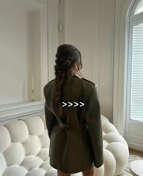 A woman from the back in an olive green coat suit, is standing in a bright room with a sofa and a window.