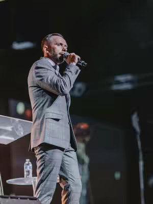 A man in a gray suit stands on a stage, holding a microphone. The background is dark with some blurry lights.