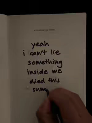 An open notebook page with handwritten text. A hand, holding a black pen, is in the bottom right corner, writing on the page. The text includes the phrases 'yeah I can't lie something inside me died this summer'.