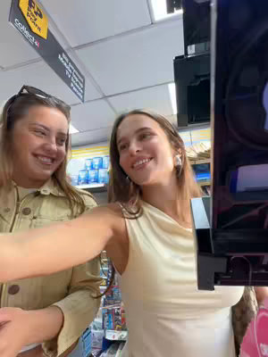 Two young women are smiling, appearing to take a selfie with a phone angled towards them. They are standing inside a store, suggesting an informal or candid moment. The lighting is bright and seems to be natural as the camera is high and the background is blurred, which suggests the use of a phone camera or similar device. The woman on the left is wearing a light-colored jacket, while the woman towards the bottom is wearing a dress, possibly in light cream color.