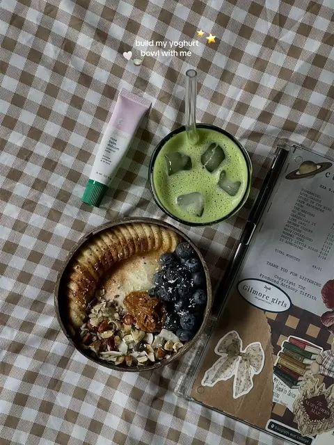 A flat lay of a breakfast scene featuring a coconut bowl filled with yogurt, banana slices, blueberries, and granola. To the side are a small container of lip balm, a glass of matcha, and a notebook. The setting is a brown plaid surface.