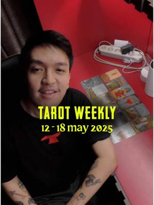 A selfie-style shot of a man smiling at the camera. He is seated at a desk with a red surface. Behind him is a red wall. On the desk, there are tarot cards and a power strip. The overlay text across the center of the image states "TAROT WEEKLY" above "12-18 may 2025".