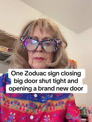 An older woman with blonde hair and large jewel-encrusted glasses is centered in the frame, looking towards the viewer. She is wearing a patterned top in shades of red and blue. Text in a white box below her face says "One Zodiac sign closing big door shut tight and opening a brand new door". The background appears to be an office space.