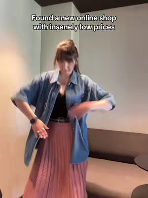 A woman in a bright room, dressed in a blue button-down shirt over a black top and a pink pleated skirt, is shown mid-action. She appears to be adjusting her outfit with a watch and rings on her wrist. Behind her, there's a background of a neutral-colored wall and a curved sofa.