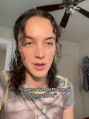 A woman with short, curly brown hair and hoop earrings is centered in the frame. She is wearing a light-colored top. The background includes a ceiling fan and part of a wall with some hanging clothes.