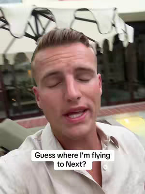 A man with light skin and short, styled blond hair faces the camera, speaking. The background features outdoor patio-like architecture with a white canopy overhead. Text on the image reads, 'Guess where I'm flying to Next?'