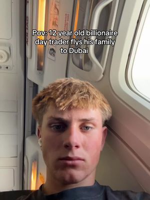 The cover image features a young man with light brown hair, positioned in the center of the frame. He's wearing a black shirt and has earbuds in. The background shows the interior of an aircraft, with a window visible to the right and part of the upper storage compartment or cabin overhead. Text overlay at the top of the image reads 'Pov: 12 year old billionaire day trader flys his family to Dubai'.