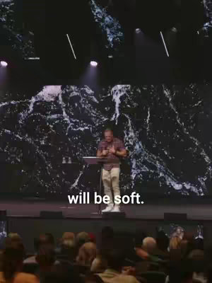 A man standing at a podium on a stage, speaking to an audience. The background is a textured pattern, and text is overlaid at the bottom. The image is softly lit, creating a warm atmosphere.