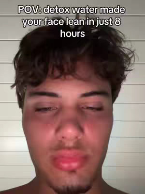 A close-up shot of a young man's face. The man has short dark hair and is looking down with his eyes nearly closed. Text overlay at the top reads, 'POV: detox water made your face lean in just 8 hours'. The background is a white, slatted wall.