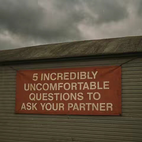 The image captures a weathered, gray building facade beneath an overcast sky. A rectangular banner, colored a rusty orange, is affixed to the building. The banner displays white, bold text: "5 INCREDIBLY UNCOMFORTABLE QUESTIONS TO ASK YOUR PARTNER."