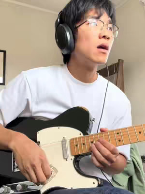 A young man with glasses, headphones, and a white t-shirt is playing an electric guitar. The background is a blurred room interior.