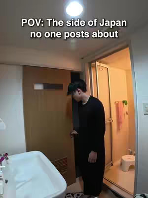 The image shows a person opening a door into a bathroom. The room is brightly lit with multiple elements: a sink, a shower area, and a small stool.