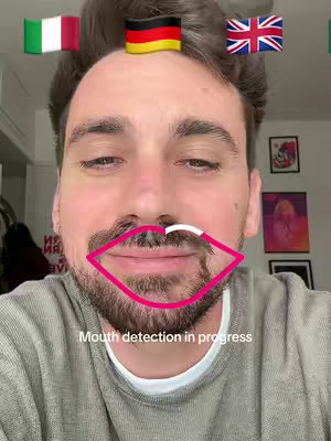 A selfie-style image showing a man's face with a filter overlay. Above his head are flag emojis of Italy, Germany, and the United Kingdom. A pink outline marks the man's lips and a white mustache is drawn on. The text overlay says 'Mouth detection in progress'.