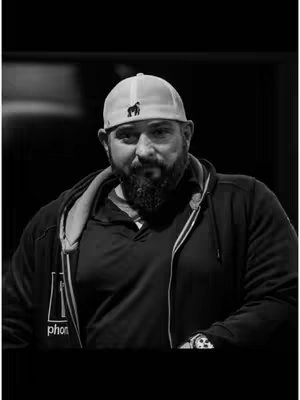 A black and white photo of a man with a beard wearing a hat and a zip-up jacket. The image is set against a dark background.