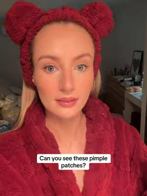 A close-up shot of a person is facing the camera. They are wearing a red fluffy headband with two large ear-like features, a red fluffy robe, and have blonde hair. The background is blurred and out of focus, with neutral tones and a hint of a dresser and other items. The main subject is staring directly at the camera, and the text overlay asks if you can see the pimple patches.