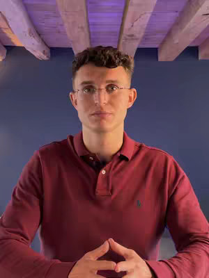 A man in a maroon polo shirt with the collar open and glasses is centered in front of a solid blue background. Above him are wooden beams creating a textured ceiling. The man's hands are clasped together, and he is making eye contact with the camera.