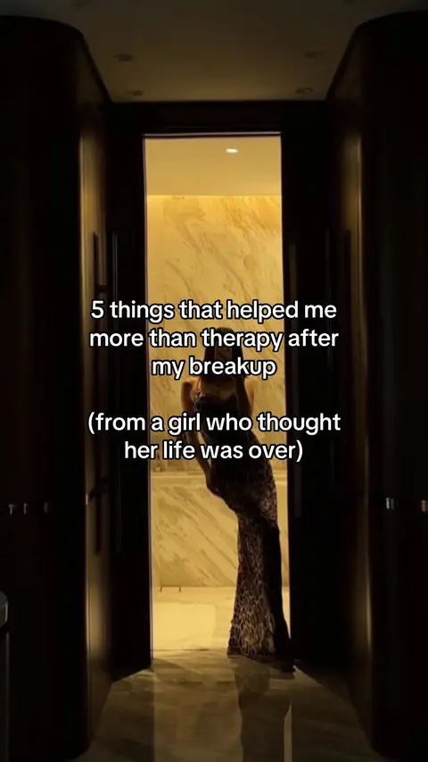 A woman stands in a well-lit doorway between two dark wooden panels. The woman is reflected in a large mirror, which further suggests she is taking a photo of herself. Light from a room beyond the door seems to shine brightly on the woman, the room filled with marble tiles, which contrast with the darkness of the hallway and the doors that encapsulate this doorway and the woman standing within.