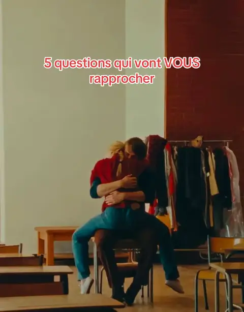 A couple hugs in a classroom. The man is seated on a chair, with the woman sitting on top of him. The man is wearing a dark shirt and brown pants, while the woman is wearing a red shirt and blue pants. The text overlay is in red and white and is at the top of the image.