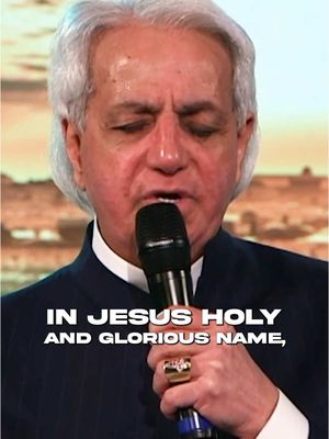 The cover image features a close-up of Benny Hinn holding a microphone to sing or speak, set against a blurred background. Text overlayed at the bottom reads "IN JESUS HOLY AND GLORIOUS NAME." Hinn is wearing a dark suit and a large ring with a prominent stone.