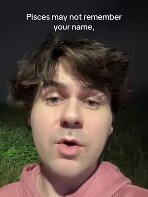 A selfie style shot of a man from the chest up, talking to the camera. The background is dark, with some green foliage visible on the lower left side. The man has short brown hair, fair skin, and is wearing a pink hoodie. Overlayed text at the top reads 'Pisces may not remember your name,' utilizing a simple, sans-serif white font.