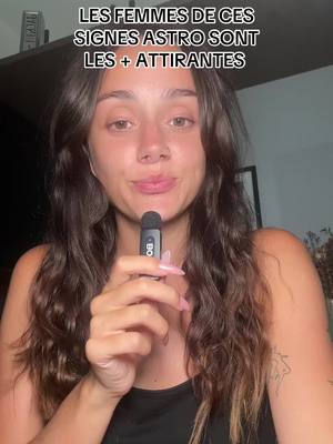 A woman with long, wavy brown hair holds a microphone and looks towards the center of the frame, facing the camera. Text overlay at the top says, 'LES FEMMES DE CES SIGNES ASTRO SONT LES + ATTIRANTES'. The background is out of focus, but shows a shelf and a picture frame.