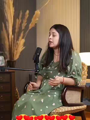 A woman is seated in a patterned chair, speaking into a microphone. She wears a green dress with white floral patterns. Behind her is a softly lit studio setup with vertical wood panels and what appears to be tall ornamental grasses.