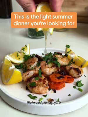 A top-down shot showcases a plate of grilled shrimp, roasted vegetables, and lemon wedges. A hand is squeezing lemon juice over the food, creating a fresh and appetizing presentation. The setup is clean, with the food arranged on a white plate against a neutral background.