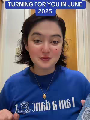 A woman with short, dark hair is centered in the frame, wearing a blue t-shirt, and looking directly at the camera. A text overlay at the top reads "TURNING FOR YOU IN JUNE 2025." She is holding a tarot deck in her right hand. The background is a light-colored wall with a wooden door frame on either side.