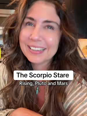 The cover image is a close-up shot of a woman with long, wavy brown hair. She is smiling with her mouth open, revealing white teeth. The background is slightly blurred hinting at an indoor setting, with a glimpse of a window or a bright light source. Superimposed on the image are two overlaid text elements. One white rectangular label is used to display "The Scorpio Stare" and below this overlay, a second horizontal label states "Rising, Pluto and Mars". The woman wears an off-white colored shirt. The overall composition is meant to be inviting and create the focal point on the woman and her expressed emotions.