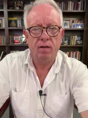 A medium shot features a man speaking directly to the camera. He has short, graying hair and is wearing glasses and a white collared shirt. The background is a bookshelf filled with books. The lighting appears to be natural, casting shadows and highlights on his face.