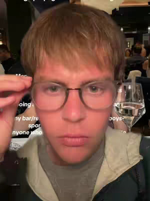 A mid-shot of a young man, with fair skin and short, light-brown hair, wearing clear-framed glasses and a gray top over a dark jacket. He looks directly at the camera with a slightly questioning expression while holding the frame of his glasses. The background is blurred but shows an indoor setting with other people present.