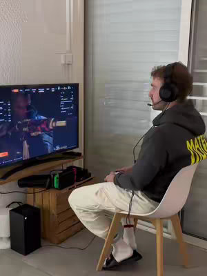 A man sits in a room in front of a large TV screen playing a video game. He wears a headset and is holding a controller. The room has a wooden crate and a window. The man is wearing a gray hoodie, beige pants, and white socks with black slides