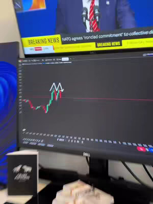 The image shows a computer monitor displaying a stock chart, with red and green lines indicating price fluctuations. Above the monitor is a television screen showing live news. The setup appears to be an office environment.
