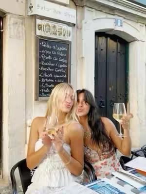 Two young women are posing for photo. One is blonde with a white dress, the other has dark hair and a floral print top. They are both holding glasses of white wine and pouting at the camera. The background is a restaurant, with a menu on a chalkboard and a dark door.