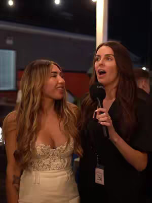 Two women are standing outdoors at night. One on the right is holding a microphone and appears to be interviewing the other. The background includes string lights and a building lit with warm lighting.