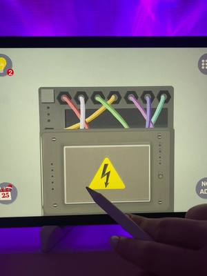 A close-up shot of a tablet screen displaying a puzzle game interface. The game features a gray box with colorful wires and a yellow warning sign. A hand holding a white stylus is in the foreground, interacting with the screen, while the background includes a purple hue.