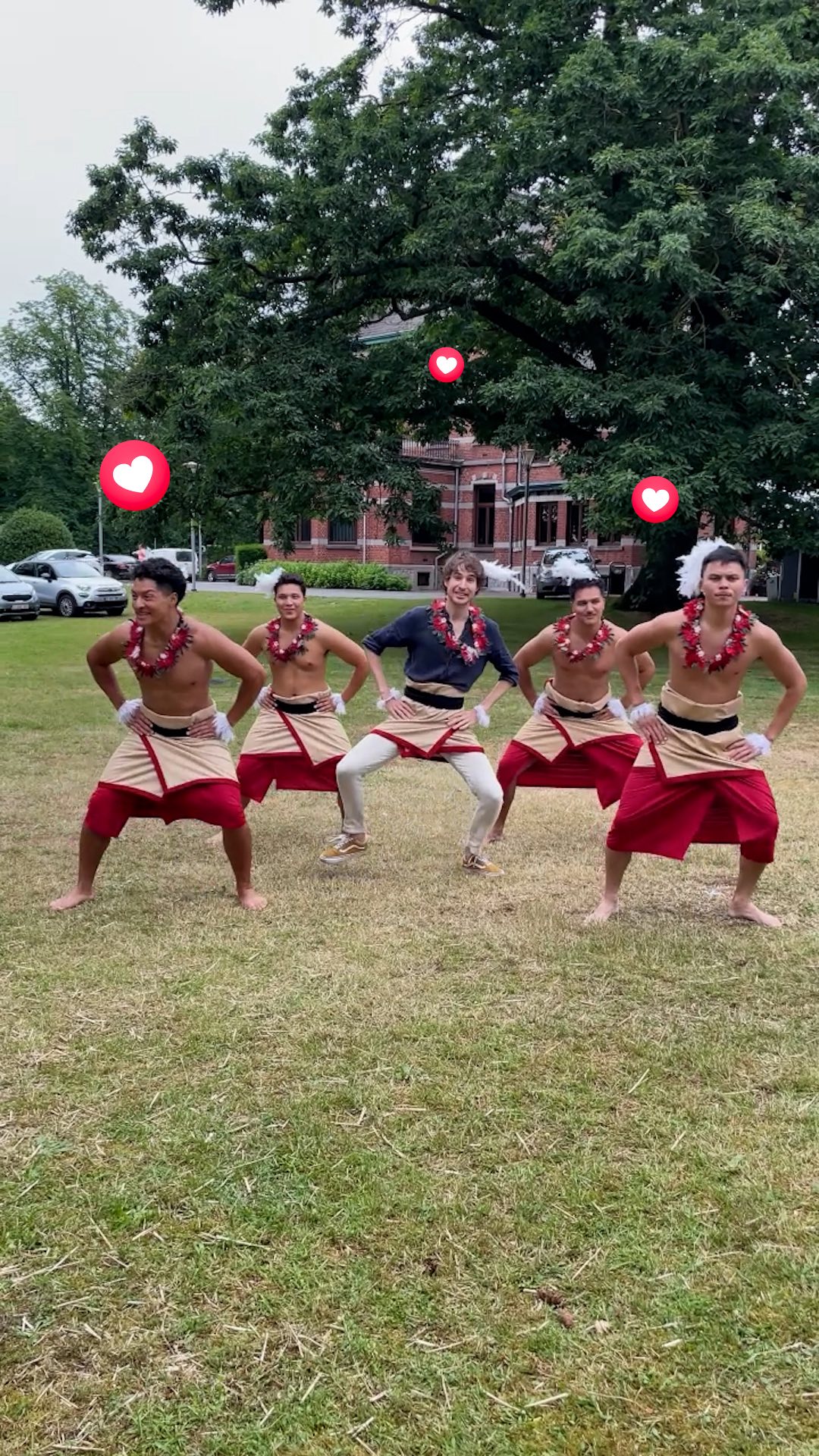 A group of five people, all appearing to be male, are posed outside in a grassy area. They are wearing traditional garments, including red and white wrap skirts and floral necklaces. The person in the center is wearing a dark collared shirt and white pants, shoes, and is posed with his hands on his waist. The other four are shirtless. The background includes a large, green tree, part of a building (possibly a large house or estate), and some cars. Floating heart emojis are added across the scene.