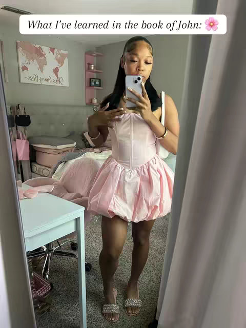 A mirror selfie in a room with pink and neutral decor. The woman is wearing a light pink dress, holding a phone up to her face, and taking a photo, with text overlayed in a bubble. The bed and a world map hangs on the wall.