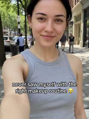 A selfie-style shot of a woman on a city street. She is in the foreground, smiling slightly. The background shows a sidewalk, some buildings, and other people walking. She is wearing a grey tank top. Text overlayed shows: 'never saw myself with the right makeup routine 🥺'