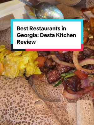 The cover image presents a close-up shot of a platter of Ethiopian food. The dish features injera bread, a spongy flatbread, topped with various foods. There are darker, cubed meats, yellow sauteed vegetables, and some fresh onion rings and tomatoes. A teal colored rectangle with white text is overlaid in the top center that states: "Best Restaurants in Georgia: Desta Kitchen Review". The background of the food shows a wooden table.