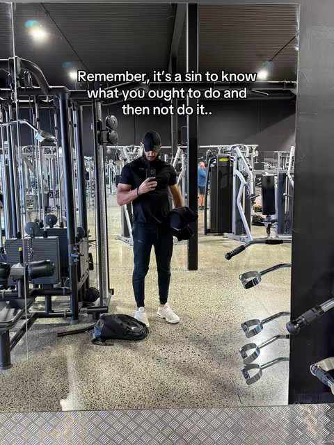A selfie taken in a gym setting, the main focus is on a man wearing a black outfit holding a phone, with text overlaid across the top that is a motivational quote. The gym environment includes various equipment, and a mirror reflects the scene.