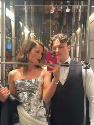 A selfie taken in an elevator with the couple facing the camera, reflecting in the mirror. The woman, on the left is wearing a shimmering, silver and white dress; The man on the right is wearing a black suit with a white shirt, and dark bow tie; He is holding a pink phone up taking a selfie.