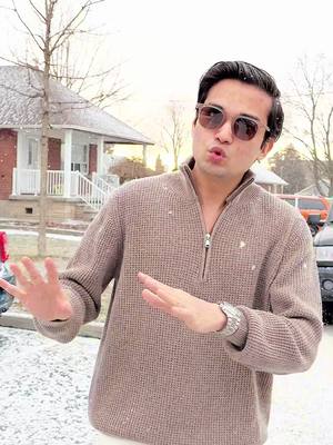 A man wearing sunglasses and a brown zip-up sweater stands in front of a house in a snowy environment, gesturing with his hands as if speaking. The image has a neutral color palette with the gray and white tones of winter.