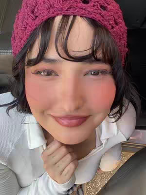 Close-up of a smiling woman is shown, angled from a slight upward position. She is wearing a maroon crocheted beanie with a fringe of dark hair peeking from underneath. Her face is made up with pink blush, and her lips are glossy and a muted pink. A glimpse of a white neckline of a shirt is visible below her chin, with hints of a textured surface in the lower portion where her hands are clasped.
