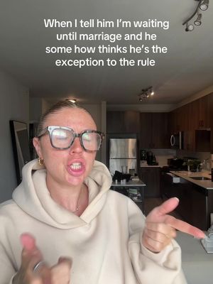 A woman with blonde hair wearing glasses and a beige hoodie speaks animatedly in front of a kitchen and living room setting. Text is overlaid at the top. She's pointing with both hands.
