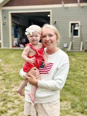 A woman holds a baby girl. The woman is fair-skinned with blonde hair, wearing a white sweater with an American flag design. The baby girl has fair skin, blonde hair, and a white bow, wearing a red floral romper. They are smiling at the camera, set outdoors with a house, grass, and trees in the background.