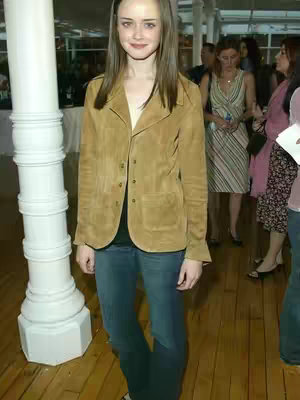 The image shows Alexis Bledel standing indoors. She is wearing a tan suede jacket over a black top and blue jeans. The background includes other people, wooden floors, and a large white pillar. The overall composition suggests an event or gathering.