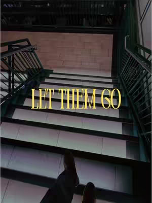 An overhead shot of a staircase. The steps are leading up, and at the bottom of the image, two feet are visible. Overlaying the staircase is the text 'LET THEM GO' in a golden color.