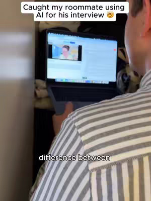 The video shows a man from behind, wearing a striped shirt, holding a laptop. The screen displays a video call with another person. Text overlays on the cover image read, 'Caught my roommate using AI for his interview'. The overall scene suggests a candid, behind-the-scenes look at someone using AI during a job interview.