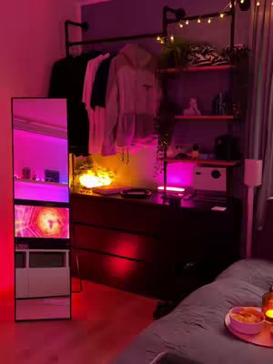 A cozy bedroom scene with a warm color palette dominated by reds and purples. A large black mirror sits in the foreground, reflecting the room and a screen displaying what looks like abstract art. The background features a black clothing rack with clothes hanging, a wooden dresser, and a bed with a tray of snacks. The lighting is soft and ambient, created by a combination of string lights and LED strips, enhancing the room's inviting atmosphere.